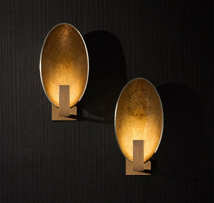 Luxury Italian oval wall sconces in brushed gold finish casting warm ambient light on a textured dark wall.
